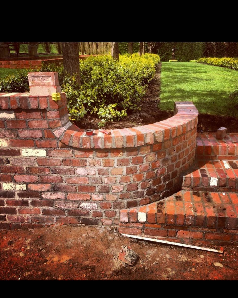 Double-tier curved red-brick planter with cascading stepped courses. Every brick cut and laid by eye.